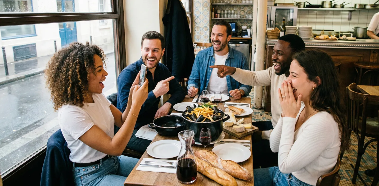 Fünf Menschen lachen an einem Restauranttisch, eine Person hält ihr Handy hoch.